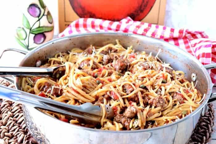 A horizontal image of a cooked pot of linguine, sausage, and capers with tongs and a red and white checked napkin in the background.