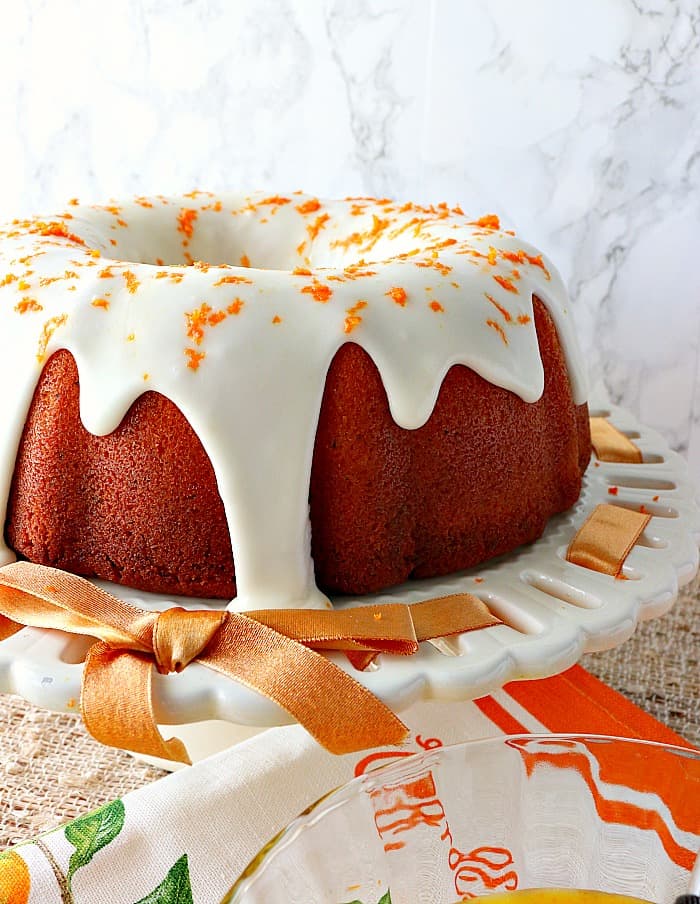 Bundt cake with a thick white icing and orange zest on a white plate with a orange ribbon.