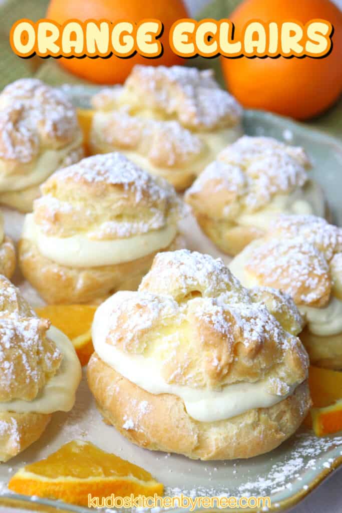 A vertical closeup of Orange Cream Eclairs on a plate topped with powdered sugar and some fresh orange slices on the plate.