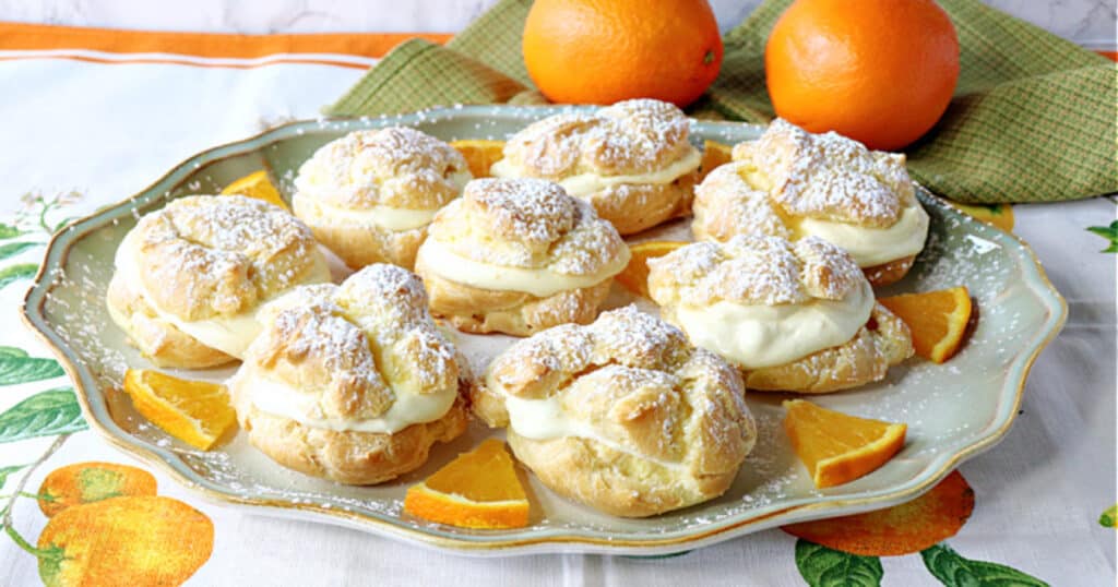 A plate filled with Orange Cream Eclairs in the foreground and two oranges and a green napkin in the background.