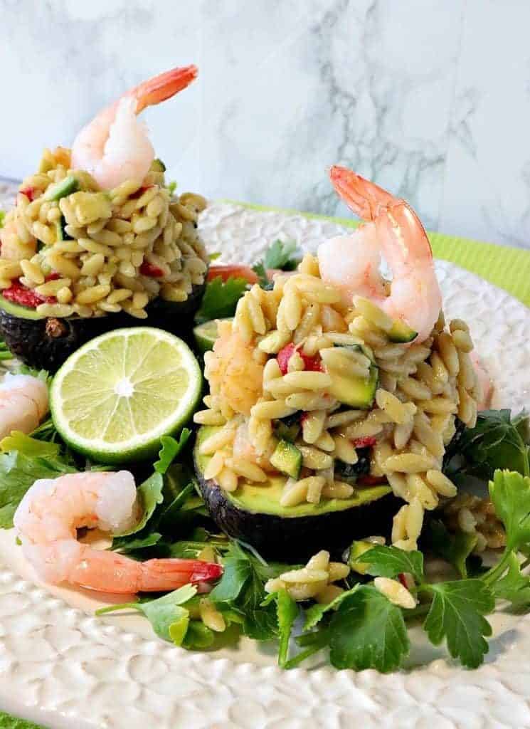 Two Orzo pasta and shrimp salad stuffed avocados with cooked shrimp on the top and a wedge of lime.