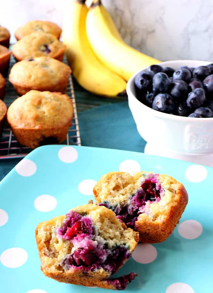 Vertical image of a blueberry banana muffin on a blue and white plate with bananas, muffins, and blueberries in the background.