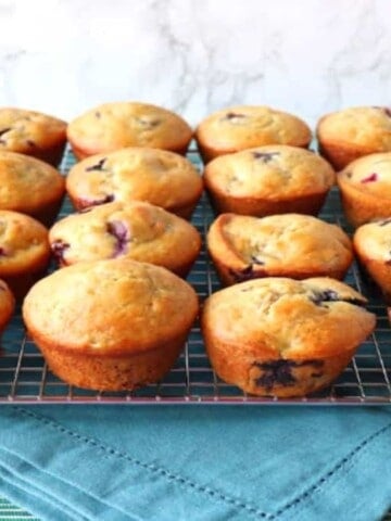 Two dozen Blueberry Banana Muffins lined up on a cooling rack with a blue napkin underneath.