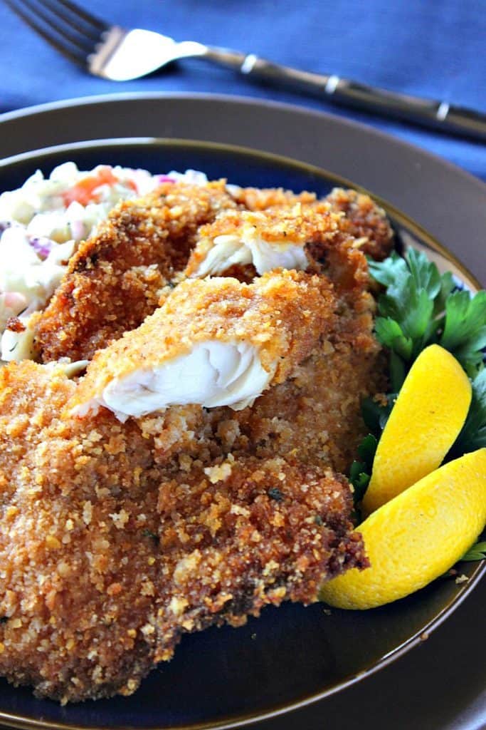 Closeup picture of flaky fried fish on a brown plate with parsley and lemon slices.