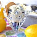 A glass dish filled with no-churn lemon blueberry ice cream with fresh lemons and vanilla cookies on the side.