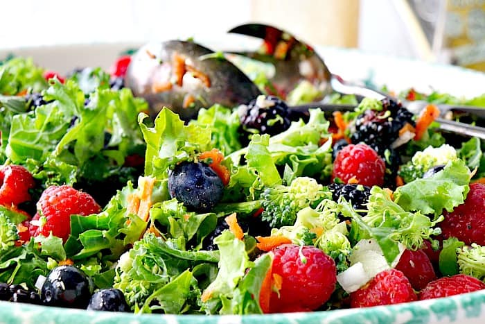 Closeup photo of a kale salad in a bowl with raspberries, blueberries, blackberries, and tongs.