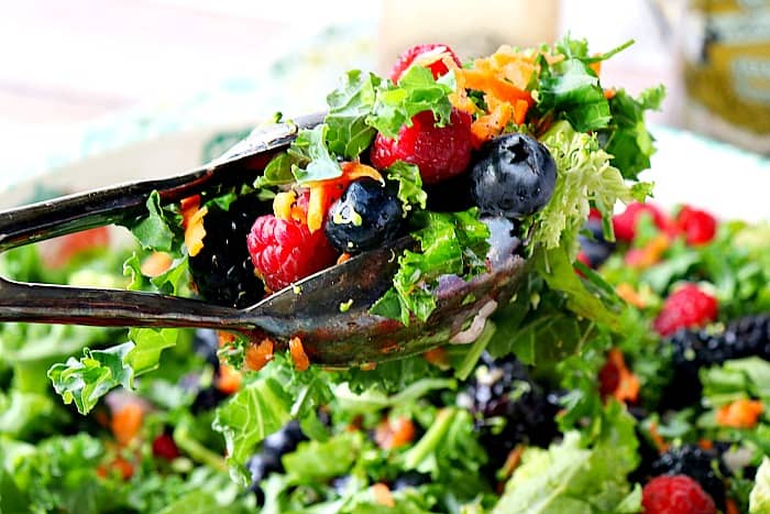 Closeup photo of salad tongs holding a kale, blueberries, raspberries, and blackberries salad over a salad bowl.