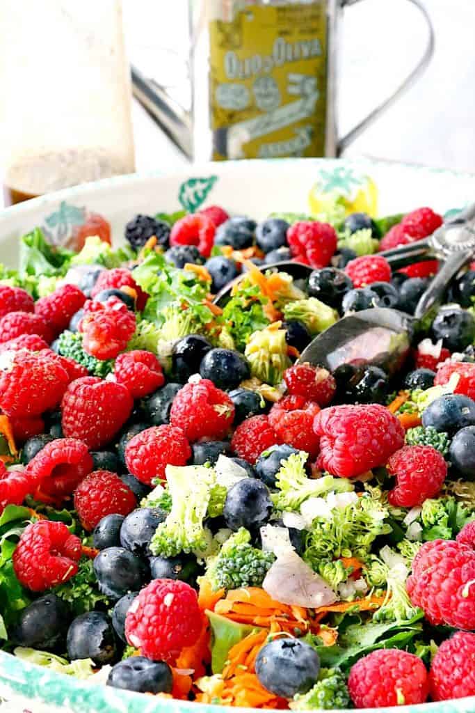 Vertical image of a kale salad with tongs, raspberries, blueberries, shredded carrots, and broccoli with a tin of olive oil in the background.