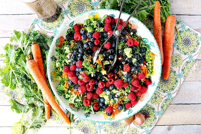 Overhead photo of a large bowl of colorful kale salad with carrots, kale, and shallots surrounding the bowl as accents. Healthy Salad Recipe Roundup.