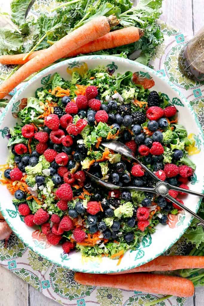 Overhead vertical photo of a large and colorful kale salad with fresh berries, carrots, broccoli and salad tongs.