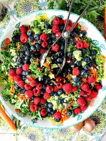 A large round bowl filled with Kale and Fresh Berries Salad and a pair of salad tongs.