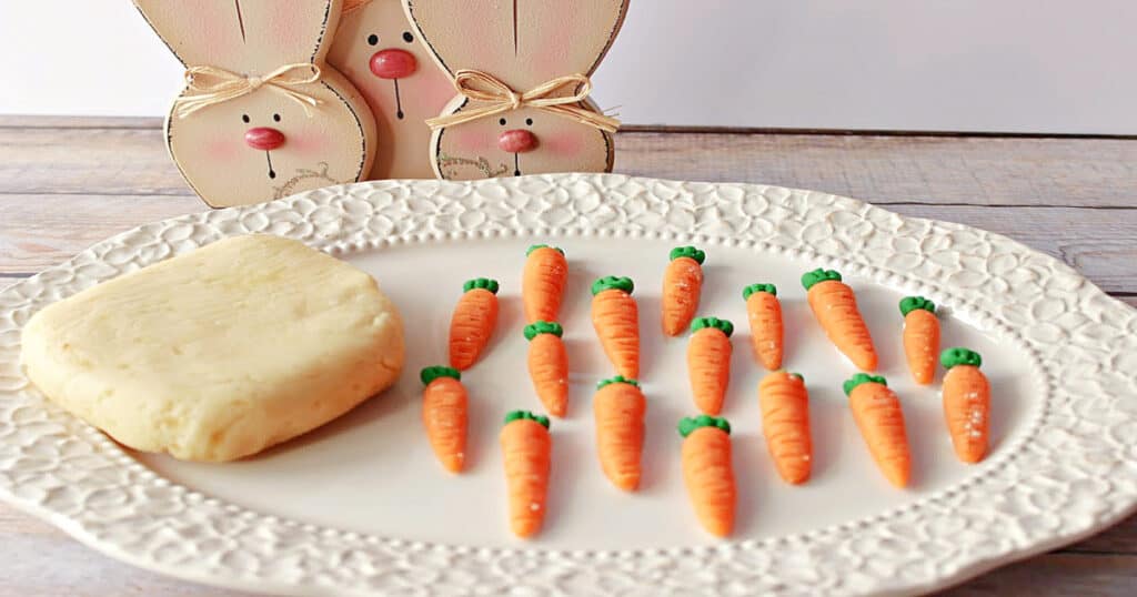 A pretty oval plate filled with Homemade Marzipan Candy Carrots with wooden bunny faces in the background.