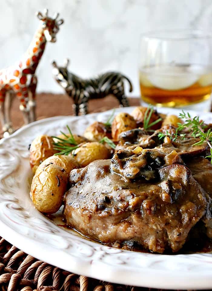 Closeup vertical image of a steak covered in mushroom sauce with a glass of bourbon and ice in the background.