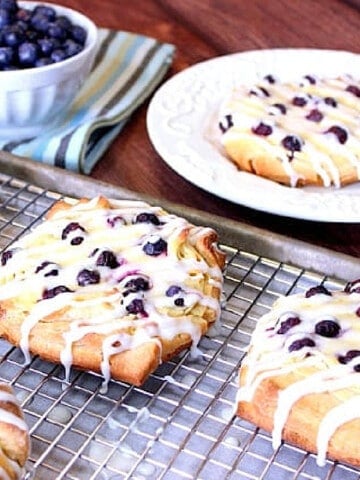 Blueberry Cheese Danish on a baking sheet with a rack with a bowl of blueberries in the background.