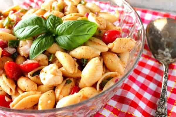 Glass bowl filled with Italian Shell Pasta Salad with fresh basil leaves and a red and white checked napkin underneath.