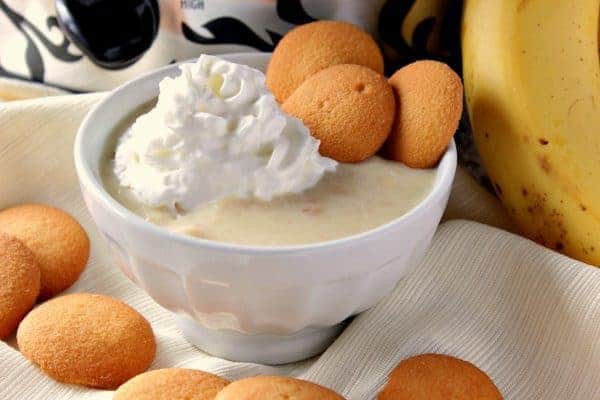 Small white bowl with banana pudding, whipped cream, and three vanilla wafer cookies. A slow cooker in the background along with bananas.
