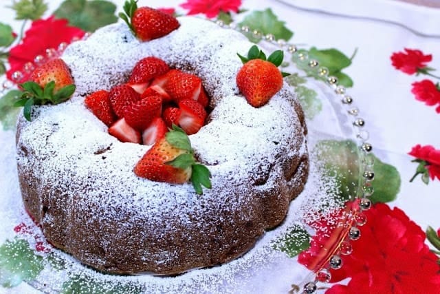 offset photo of a strawberry rhubarb bundt cake on a glass platter with confectioners sugar and fresh strawberries.