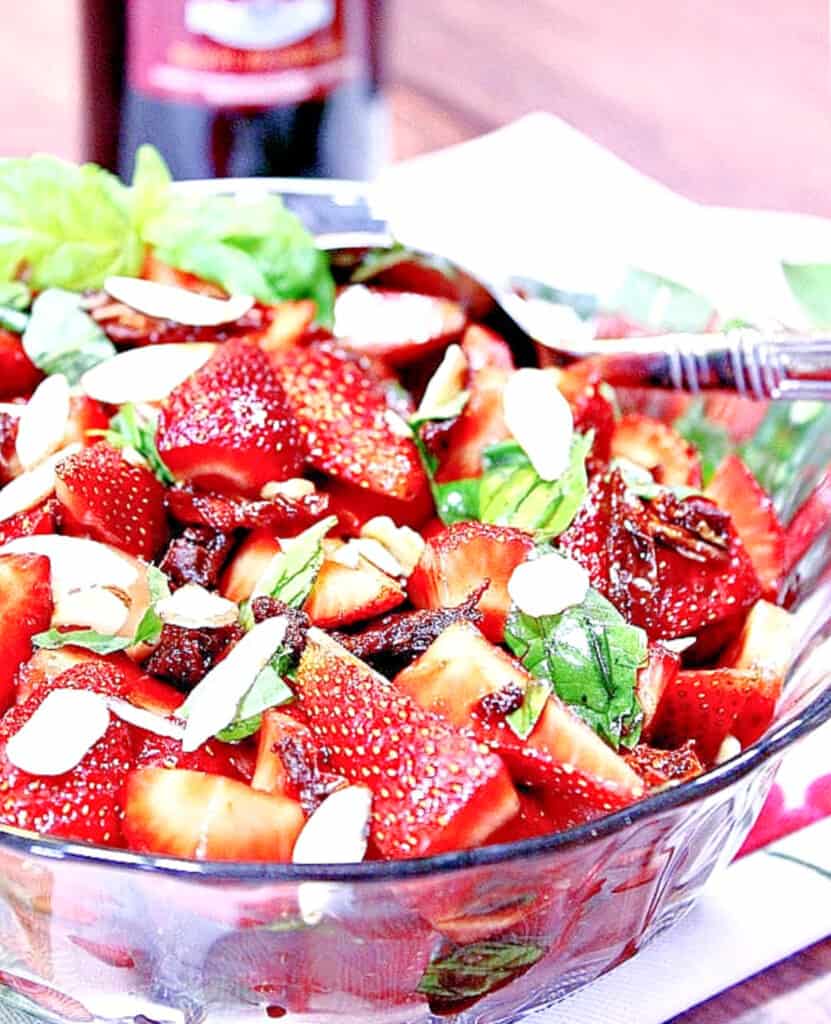 A vertical closeup photo of a Strawberry Basil Salad in a glass bowl with sun dried tomatoes and almond slices.