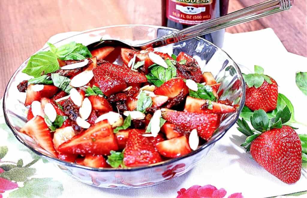 A red, green, and white Strawberry Basil Salad in a glass bowl with basil, almonds, and sun dried tomatoes.