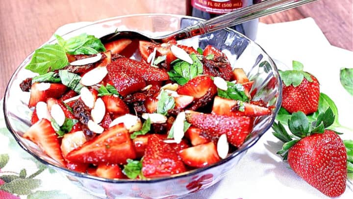 A red, green, and white Strawberry Basil Salad in a glass bowl with basil, almonds, and sun dried tomatoes.