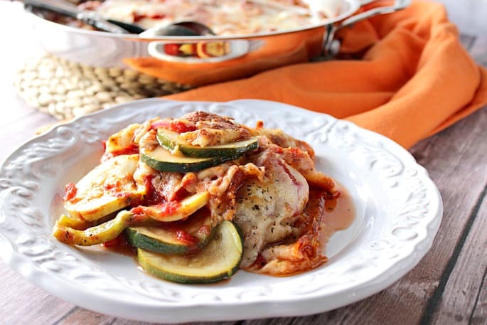 A white scalloped plate in the foreground filled with zucchini Parmesan side dish along with an orange napkin and a skillet in the background.