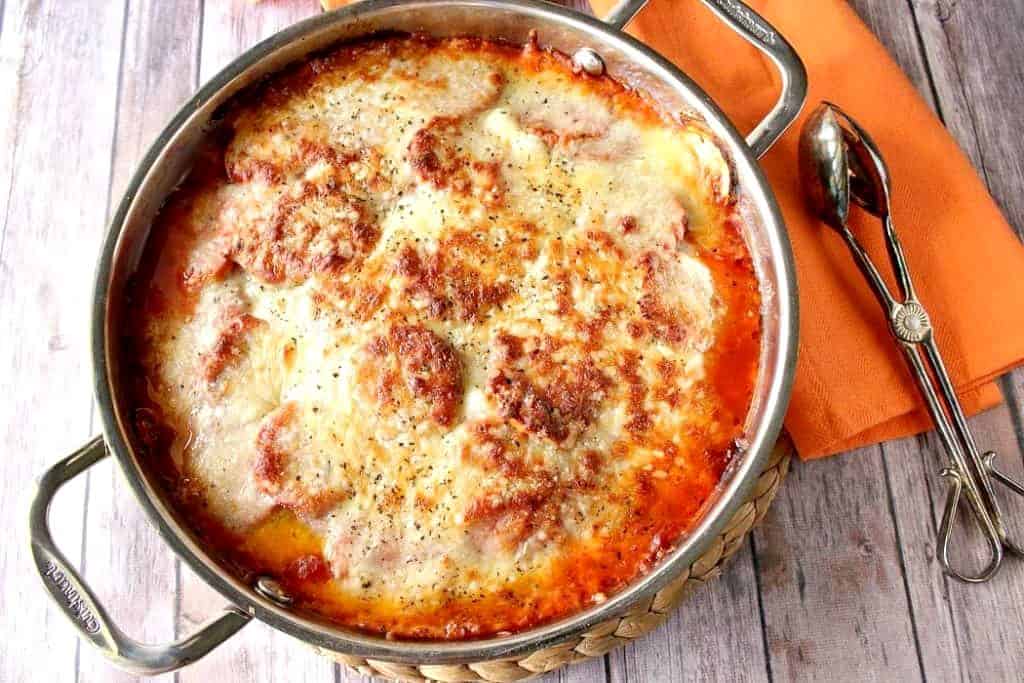 Overhead photo of a silver skillet filled with Italian zucchini Parmesan with an orange napkin and serving tongs.