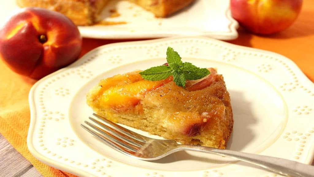 A slice of nectarine upside down cake on a white plate with a fork and a sprig of mint for garnish