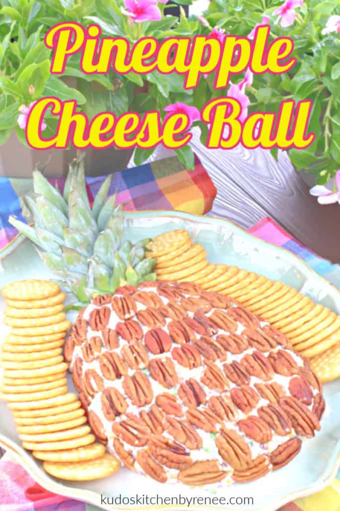 A pineapple cheese ball with crackers on the side and flowers in the background with a title text overlay graphic