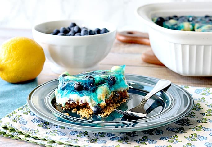 A horizontal photo of a slice of blueberry pretzel dessert on a blue plate with a fork and a lemon in the background.