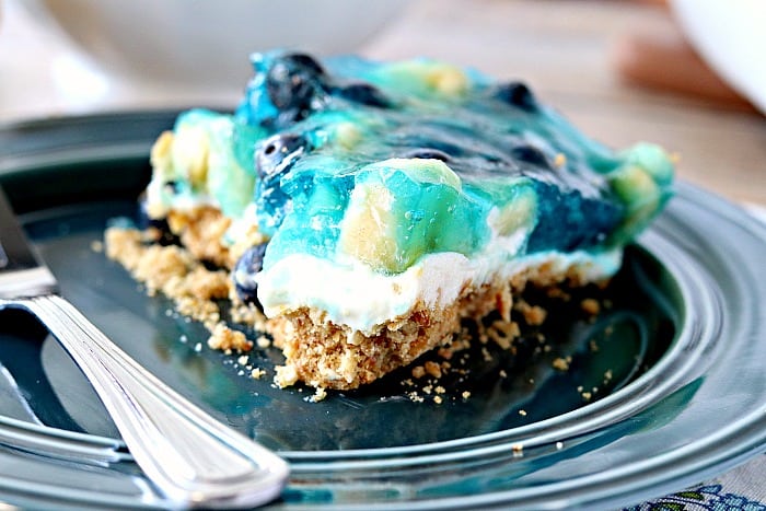 Closeup photo of a slice of blueberry pretzel dessert with bananas, blue jello, pretzel crust, and cheesecake layer on a blue plate.