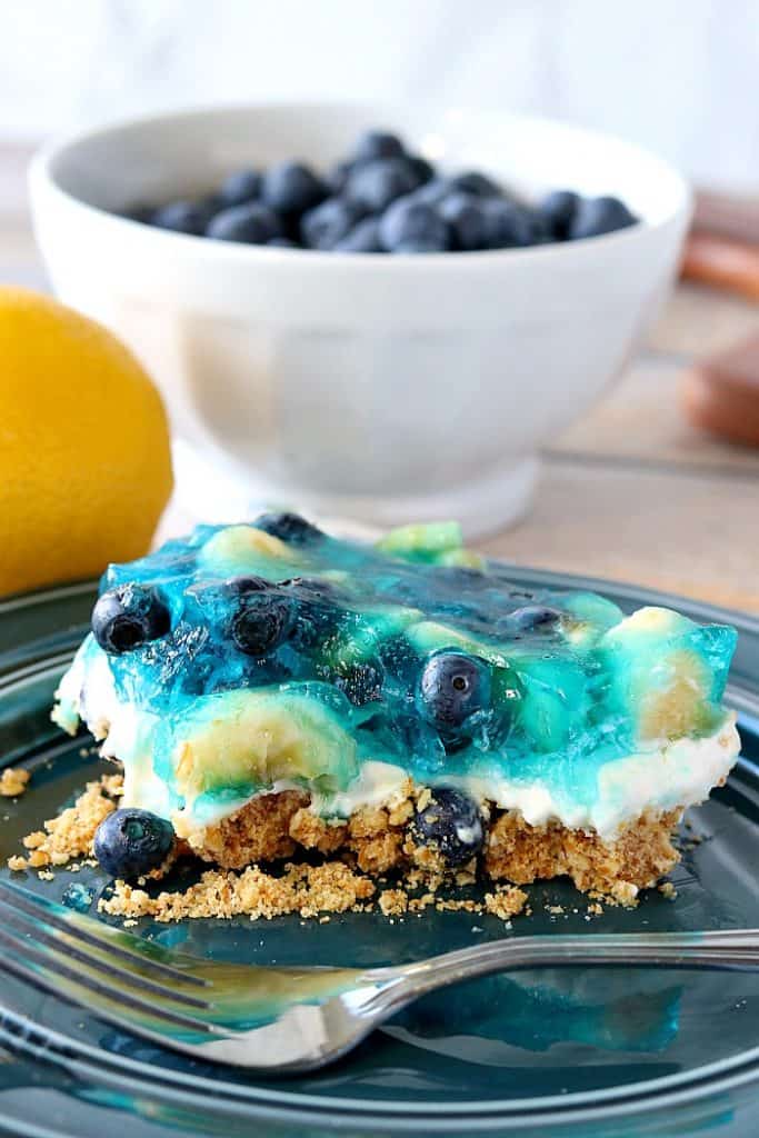 A vertical closeup photo of a blueberry pretzel dessert on a blue plate with a fork and a bowl of blueberries in the background.