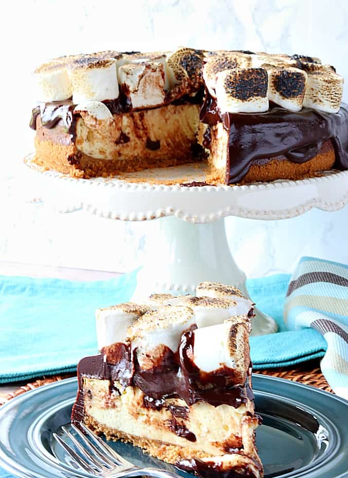 A slice of chocolate chip s'mores cheesecake on a plate with a cake stand with the cheesecake in the background.