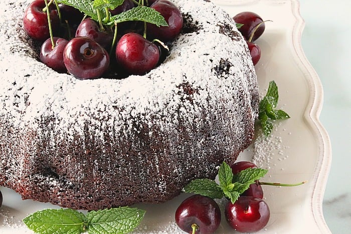 Closeup photo of a chocolate cherry bundt cake with a confectioners sugar dusting, fresh cherries, and mint.