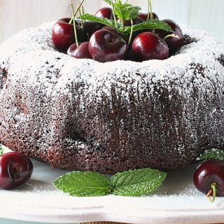A chocolate bundt cake on a white plate with cherries and mint.