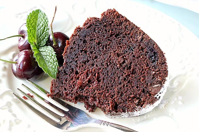A slice of chocolate cherry bundt cake on a white plate with a fork, cherries, and mint.