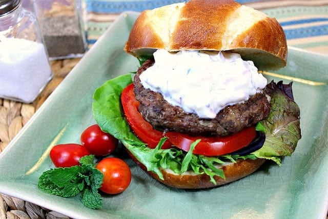 Horizontal photo of a Greek lamb burger on a square aqua color plate with tzatziki sauce and tomatoes.