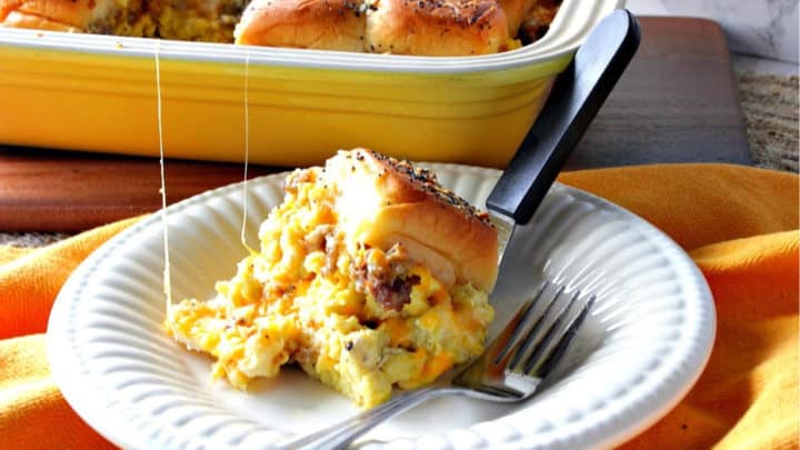A scrambled egg breakfast slider on a plate with a fork and a casserole dish filled with sliders are in the background