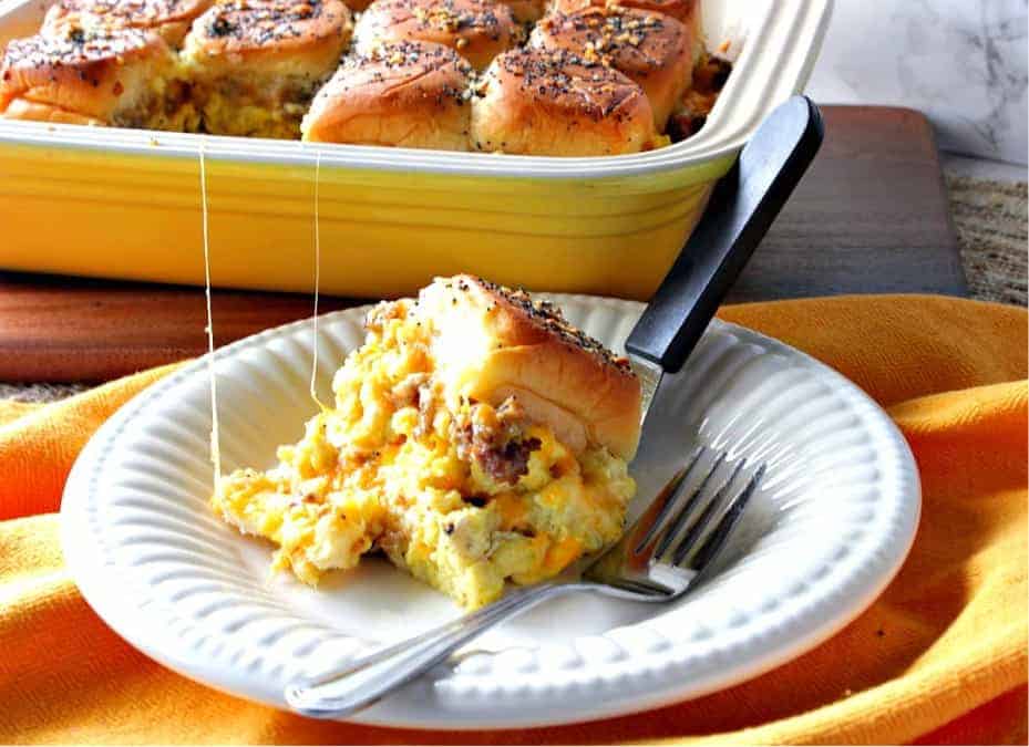 A scrambled egg breakfast slider on a plate with a fork and a casserole dish filled with sliders are in the background