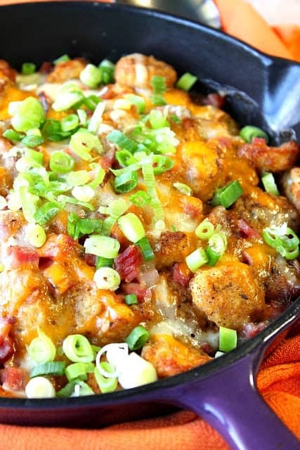 Closeup photo of a skillet loaded with tater tot poutine, cheese, ham, and green onions.
