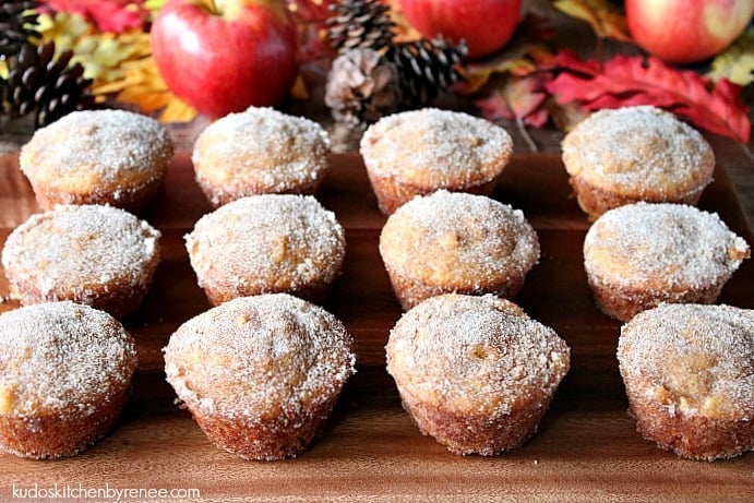 One dozen Apple Cider Donut Muffins on a wooden board with apples and autumn leaves in the background.