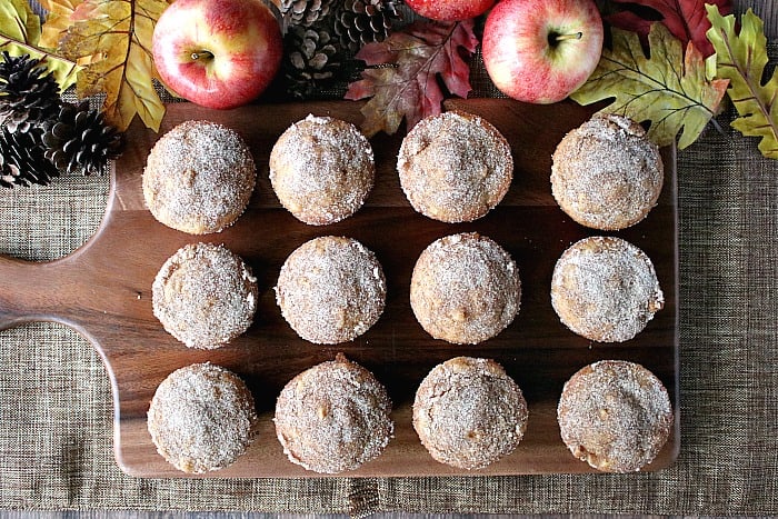 Overhead photo of a dozen Apple Cider Donut Muffins on a wooden board with apples, acorns and autumn leaves.