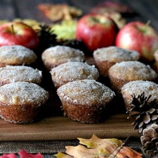 A bunch of apple cider donut muffins on a wooden board with autumn leaves and apples in the background.