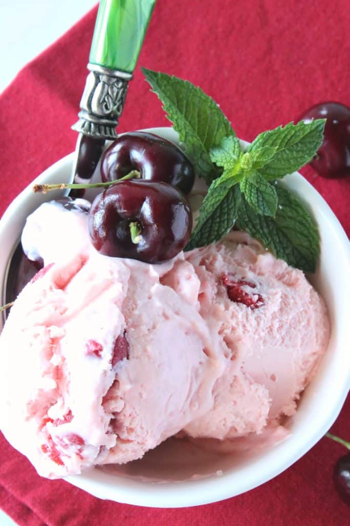 Closeup of a bowl of cherry chunk amaretto ice cream with a green spoon and a sprig of mint.
