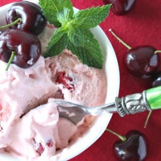 Offset photo of a dish of cherry chunk amaretto ice cream on a red tablecloth with a green spoon and a sprig of mint.