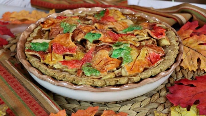 A colorful autumn leaves apple pie on a rattan place mat surrounded by fall leaves.