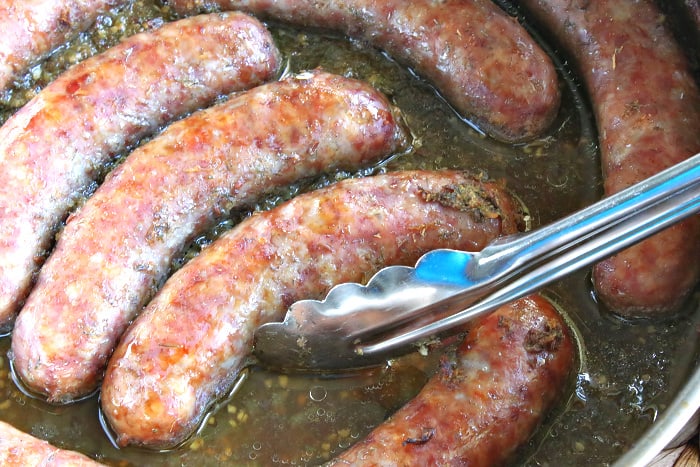 Closeup overhead photo of bratwurst in beer with dill weed and a pair on tongs.