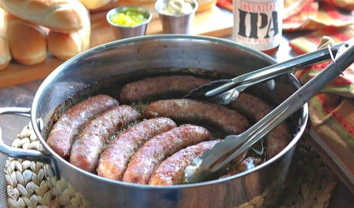 A large skillet of beer braised bratwurst with a pair of tongs. Buns and beer are in the background.