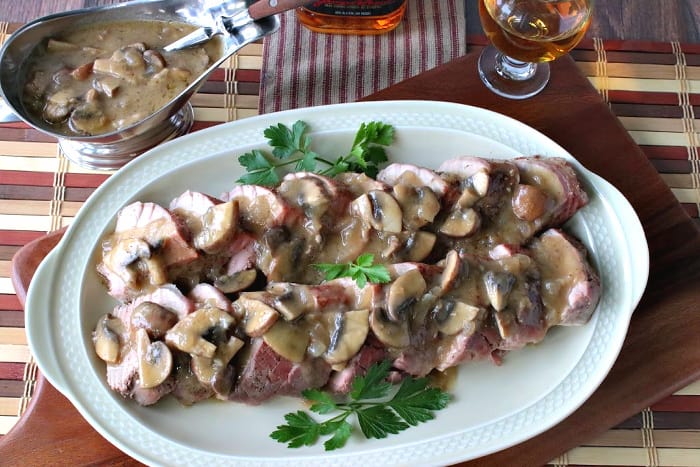 Overhead photo of a pork tenderloin on an oval platter covered with mushroom gravy, with a gravy boat and a glass of bourbon.