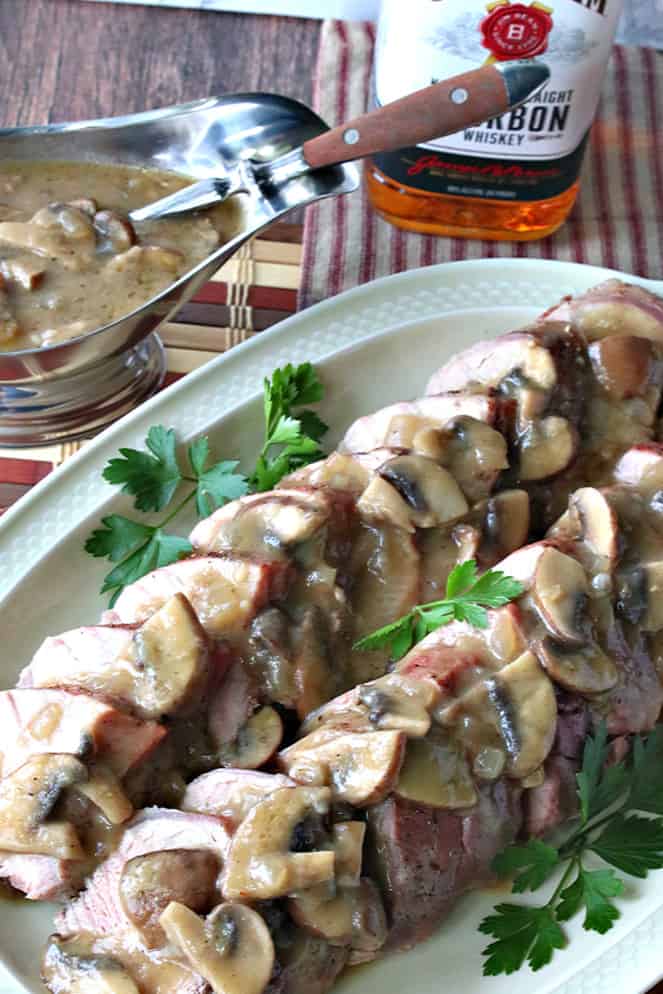 A vertical photo of a pork tenderloin with mushroom gravy on a platter with parsley and a gravy boat.
