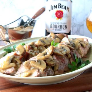 A sliced pork tenderloin on a platter with mushroom gravy and a glass of bourbon in the background.
